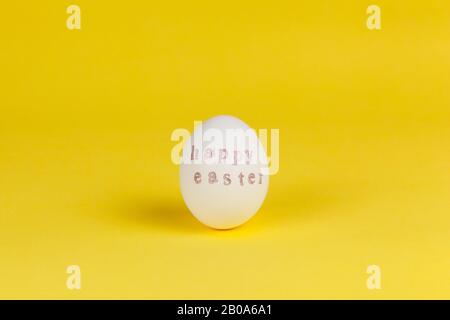 One white chicken egg with words Happy Easter written with help of stamps and pink metallic paint on yellow background, copy space. DIY, festive conce Stock Photo