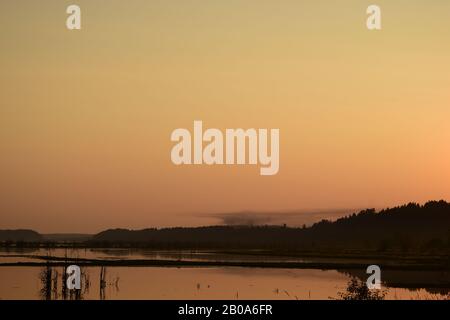 Silhouetted Trees By Large Body Of Water During Wildfire Against Beautiful Sunrise Sky And Smoke Stock Photo