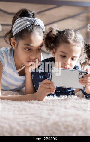 Two dark-skinned young girls, sisters, are lying in bed and looking at a smartphone against the background of the sun from the window. Persian girls Stock Photo
