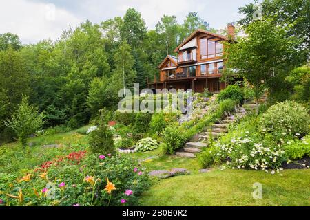 Rear of golden-brown stained piece sur piece Eastern white pine log and timber home and sloped backyard garden with cascading waterfall and stream. Stock Photo