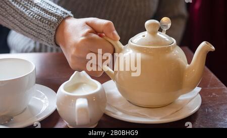 Woman's hand holding a traditional white English teapot. Close-up of girl's hand picking up a full pot of tea with empty cup and saucer, and milk jug. Stock Photo