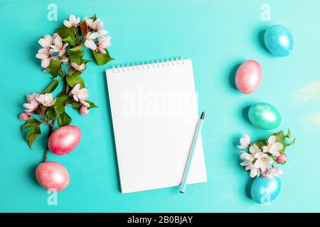 Festive Easter layout with pink Apple tree twigs, colored Easter eggs, white blank notepad with pen on turquoise background. Easter composition. Top v Stock Photo