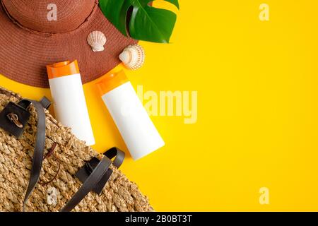 Sunscreen lotion bottles, green tropical leaves, rattan bag and female beach hat on yellow background. Sun protection, suntan cosmetic product, summer Stock Photo