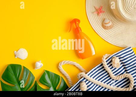 Orange sunscreen spray bottle, tropical leaves, striped beach bag and female hat on yellow background. Sun protection, suntan cosmetic product, skin c Stock Photo