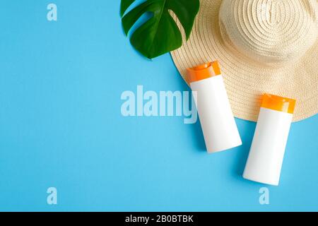 Sunscreen lotion bottles, green tropical leaf, female beach hat isolated on blue background. Sun protection, suntan cosmetics, skin care concept. Flat Stock Photo
