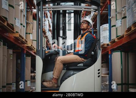 Happy forklift driver focused on carefully transporting stock from shelves around the floor of a large warehouse wearing a white helmet and vest Stock Photo