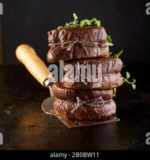 Gourmet thick juicy grilled fillet steak medallions stacked together on a metal spatula and garnished with fresh herbs in square format side on agains Stock Photo