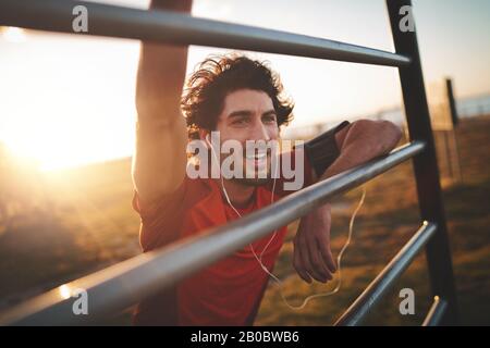Portrait of a cheerful young handsome male runner with earphones in his ears leaning on railing after exercising in the park on a sunny day Stock Photo