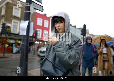 London, England, UK. Young Japanese woman using her mobile phone in Camden on a rainy day Stock Photo