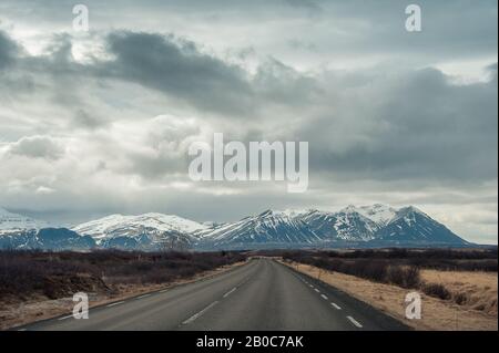 Empty road leads toward a snow capped mountain with moody, cloud sky. Stock Photo