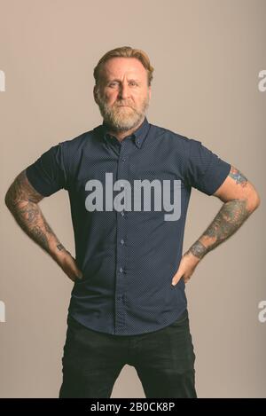 Studio shot of mature bearded man with hand tattoos posing with hands on hips in gray background Stock Photo