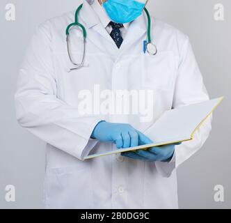 male doctor in a white coat with buttons, wearing blue sterile gloves, holding a paper open notebook in his hand on a white studio background Stock Photo