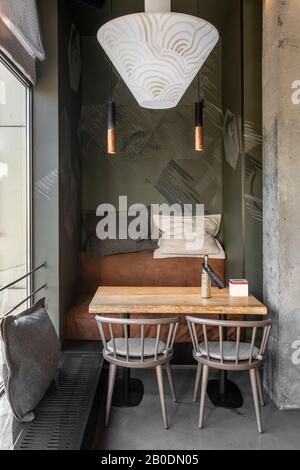 Cafe in a loft style with textured walls and concrete column. There is a wooden table with a sofa and chairs, pillows, window with a curtain, hanging Stock Photo