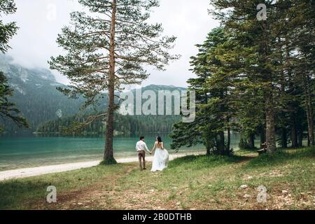 Young beautiful couple of newlyweds hold hands and walk by the lake during their honeymoon or they just got married Stock Photo