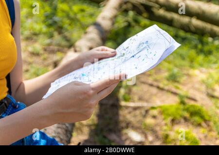 Young girl tourist with a blue backpack holds a route map sitting on a log in the forest. Autumn tourism concept Stock Photo