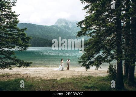 Young beautiful couple of newlyweds hold hands and walk by the lake during their honeymoon or they just got married Stock Photo