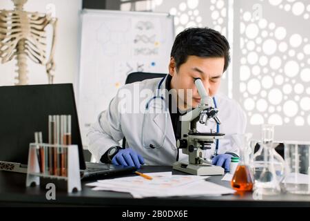 Shot of a Korean young man scientist looking through the lens of a microscope while sitting at the table with laptop, test tubes and flasks. Science Stock Photo