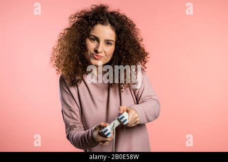 Pretty young girl playing video exciting game on Tv with joystick on pink studio wall. Using modern technology. Stock Photo