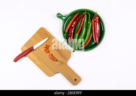 Green and red hot chilli peppers on green plate, sliced pepper on a wooden board with knife, white background, chopping board, flat lay. Stock Photo