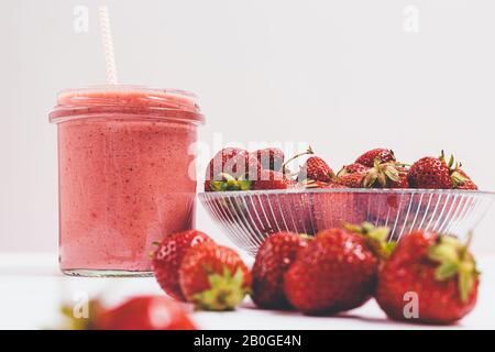 Fresh strawberries plate next to smoothie in glass jar with straw on white background, close-up. Stock Photo