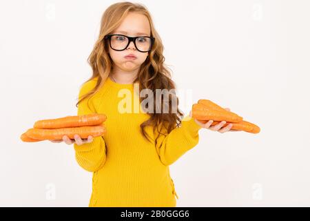 Boring teenager girl with red hair, hoody and yellow trousers holds carrot isolated on white background Stock Photo