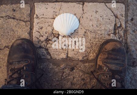 The way of saint James.well worn Walking shoes on flagstones  with scallop shell, marker for compostelle way walk. Stock Photo