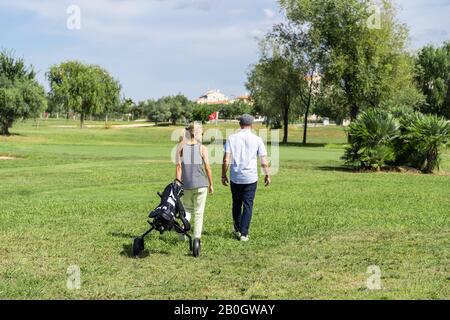 Young couple walking on a golf course with a cart to transport the equipment with buildings and trees in the background Stock Photo