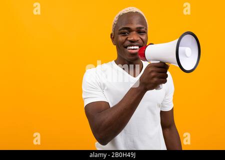 black handsome smiling american man in white t-shirt speaks news through a megaphone on isolated orange background Stock Photo