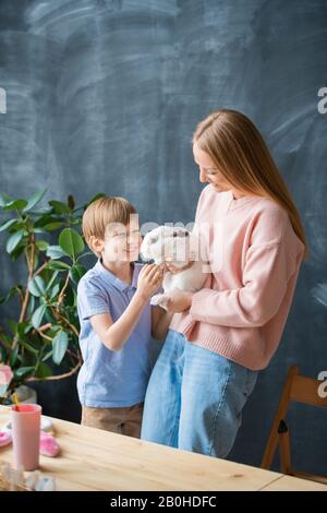 Excited cute son stroking rabbit held by mother against blackboard, Easter celebration concept Stock Photo