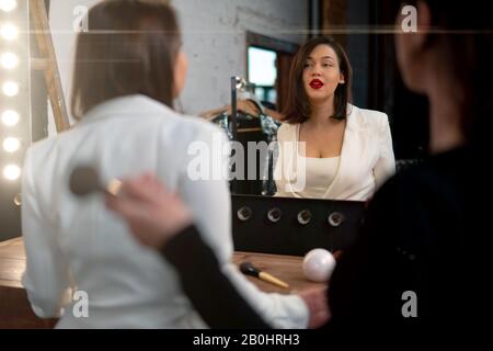 Attractive Beautiful Woman Singer Looking at Her Reflection In Big Illuminated Mirror After Make Up. Super Star Getting Ready For Performance Stock Photo