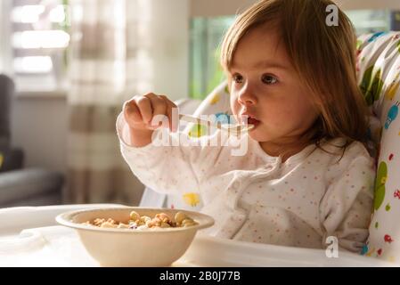 Two years old eats brakefast by herself with a spoon. Stock Photo