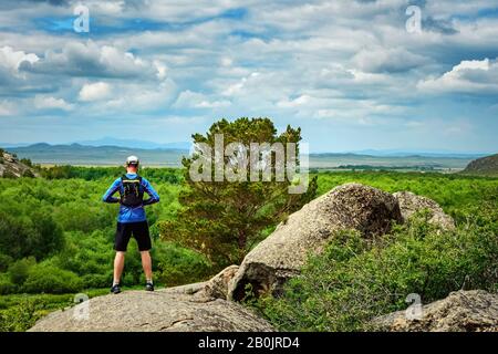 Trail runner athlete stands on the edge of a cliff overlooking a magnificent landscape. The view from the back. Man in blue jersey and black shorts tr Stock Photo