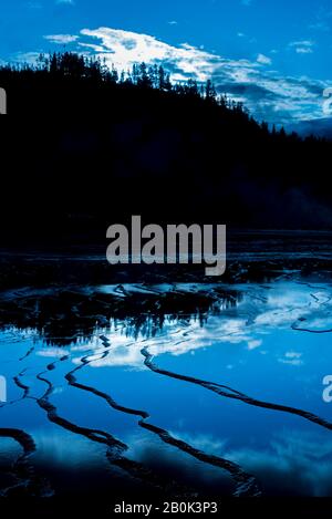 Textured mirror like rock surface reflecting the trees and sky. Forested hills under blue skies with white clouds. Stock Photo