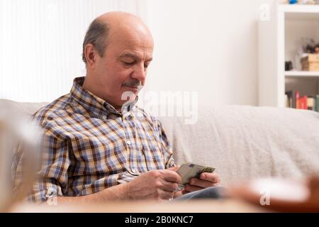 Senior bald man with mustache busy counting money and bills at home, sitting at home alone. Stock Photo