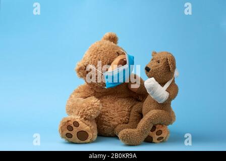 big brown teddy bear sits in a medical mask, in his hands he holds a small toy of a bear with a bandaged white bandage paw, blue background Stock Photo
