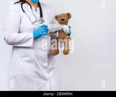 female medic in a white coat with buttons holding a brown teddy bear and bandaging her paw with a white gauze bandage, concept of pediatrics and anima Stock Photo