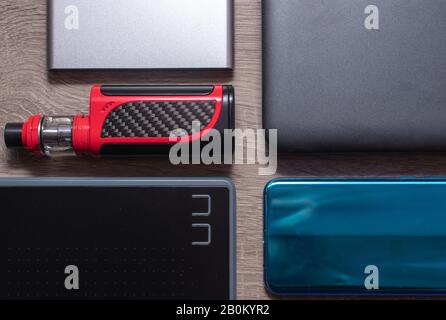 Modern technology flat lay on a grey wooden table. Silver powerbank , red vaping device , black graphics tablet, blue smartphone, grey laptop Stock Photo