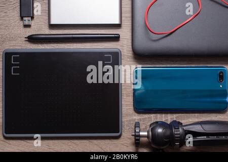 Modern technology and photography flat lay on grey wood table. Blue smartphone, grey laptop with red audio cable, graphics tablet, camera tripod, usb Stock Photo
