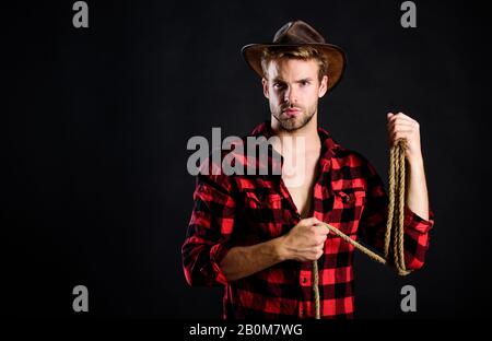 Lasso tool of American cowboy. Still used on ranches to capture cattle or other livestock. Western life. Man unshaven cowboy black background. Cowboy wearing hat hold rope. Lassoing on prairie. Stock Photo