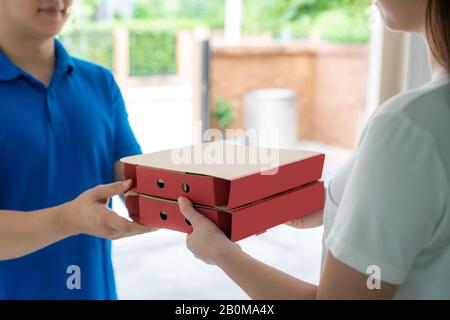 Asian delivery young man in blue uniform smile and holding pizza boxes in front house and Asian woman accepting a delivery of pizza boxes from deliver Stock Photo