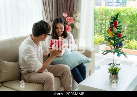 Asian man give the woman a red gift box in which there is a bottle of perfume. The woman was picked up on the sofa at home. The Christmas tree into th Stock Photo
