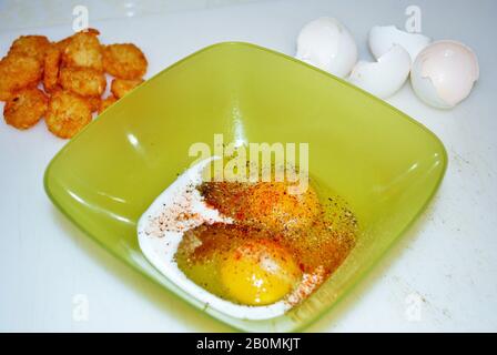 Close up of two raw seasoned eggs in a green plastic bowl hash browns and eggshells Stock Photo