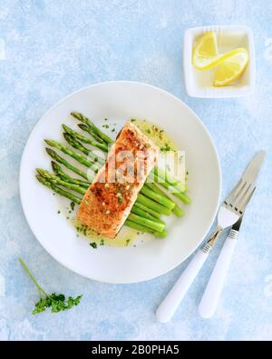 Fresh grilled wild coho salmon filet with lemon butter sauce on a bed of steamed organic asparagus. White dishes on pale blue background. Healthy eati Stock Photo