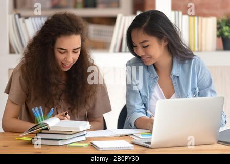 Two smiling diverse Asian and Caucasian girls students studying together, preparing exam in library with books and laptop, college university friends Stock Photo