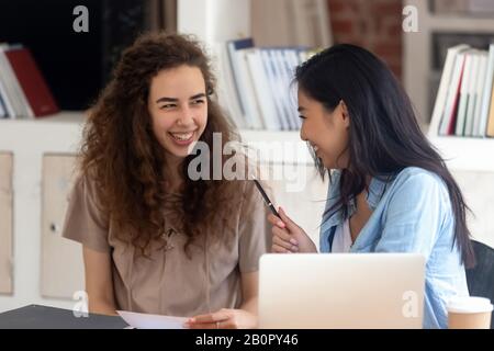 Two laughing diverse girls students studying together in library, having fun during break, talking, gossiping, Asian and Caucasian college friends dis Stock Photo