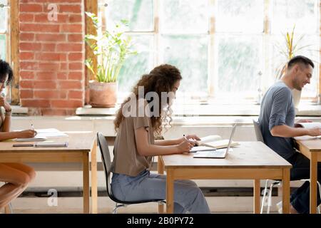 Diverse university college undergraduate students sitting at desks in row, multinational classmates writing final or admission exam in classroom, stud Stock Photo