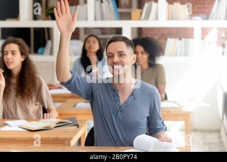 Smiling university college student raising hand, asking question during class, attractive man sitting at desk in classroom with multinational diverse Stock Photo