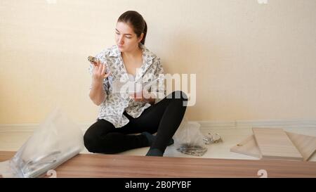 serious housewife sits on floor and reads directive looking at cabinet furniture fasteners in apartment kitchen closeup Stock Photo