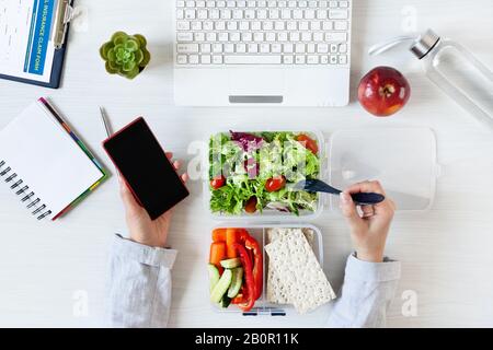 Hands of business woman working on phone and eating healthy business lunch at workplace. Vegetables and fresh green salad in lunch box on working desk Stock Photo