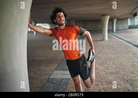 Portrait of a smiling fit young man enjoying music on his earphone warming up before running under the bridge Stock Photo